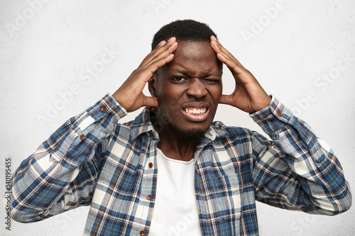 Indoor shot of frustrated annoyed dark-skinned student having bad headache, squeezing his temples, looking at camera with painful expression, can't stand upcoming college exams stress and tension