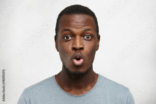 Close up shot of funny young African American man dressed casually pouting lips and raising brows, expressing shock or surprise, posing against white wall background with copy space for your text