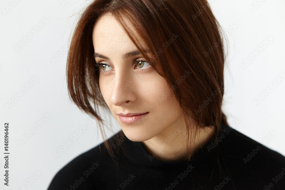 Studio shot of attractive European model with dark hair having dreamy and thoughtful expression on her face, looking sideways with faint smile, posing against white wall background wearing black dress Studio shot of attractive European model with dark hair having dreamy and thoughtful expression on her face, looking sideways with faint smile, posing against white wall background wearing black dress