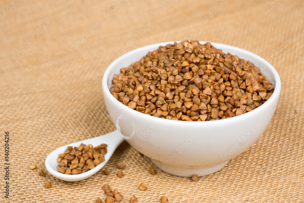 dry buckwheat heap in white bowl with spoon over sackcloth dry buckwheat heap in white bowl with spoon over sackcloth