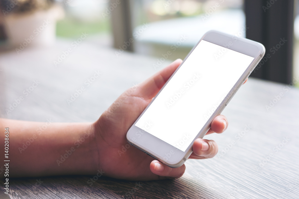 Mockup image of hand holding white mobile phone with blank white screen in vintage wooden table in cafe