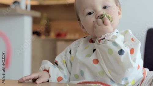 Eleven months old baby girl sitting by the desk, eating food with her fingers; baby led weaning; concept of family life and parenting
