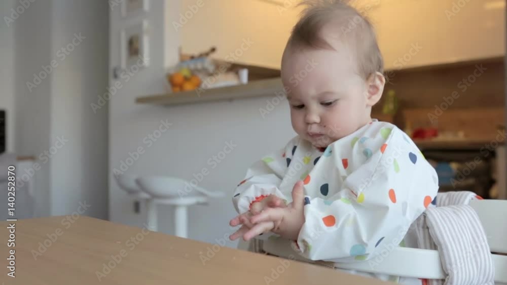 Eleven months old baby girl sitting by the desk, eating food with her fingers and then from the table top; baby led weaning; concept of family life and parenting Eleven months old baby girl sitting by the desk, eating food with her fingers and then from the table top; baby led weaning; concept of family life and parenting