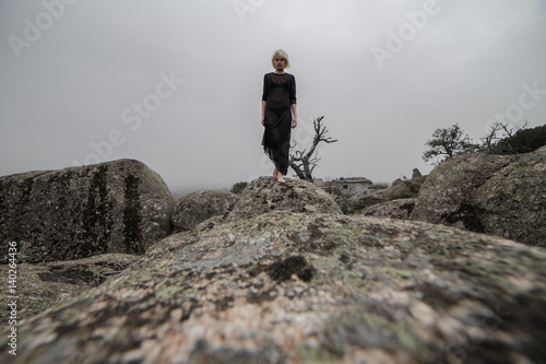 Artistic photo of a blond girl in a transparent black dress walking on the rocks in the field a cloudy day. The aesthetic is sinister and a little dark. 
