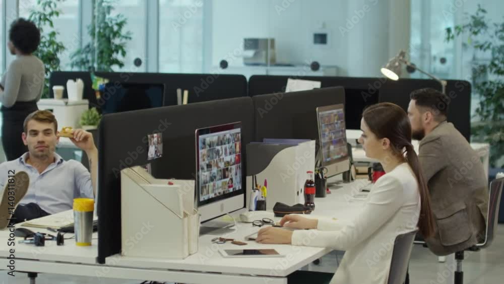 Tracking shot of modern comfortable office: one man eating pizza in workplace and putting feet on table; office workers surfing the Net on computers and two businesswomen speaking in the background Tracking shot of modern comfortable office: one man eating pizza in workplace and putting feet on table; office workers surfing the Net on computers and two businesswomen speaking in the background