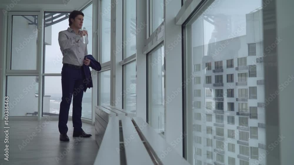 handsome young businessman have a break at work drinking water and looking on the street with urban landscape and high rise buildings. successful employer holding bottle with clear drink and relaxing. handsome young businessman have a break at work drinking water and looking on the street with urban landscape and high rise buildings. successful employer holding bottle with clear drink and relaxing.