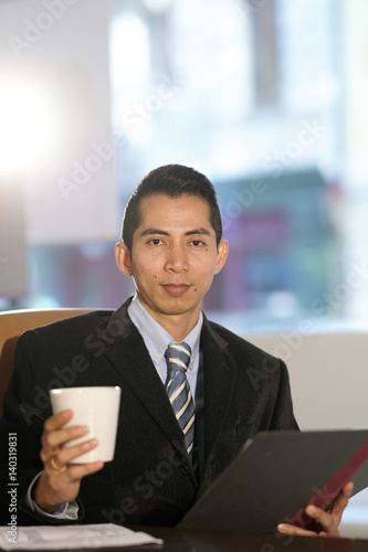 Asian manager posing at desk in office holding clipboard and tea mug. Vertical shot