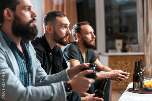 Men with a beard sitting on the couch at home with beer and chips with joysticks in hand playing computer video games. The concept of friendship, technology and weekend