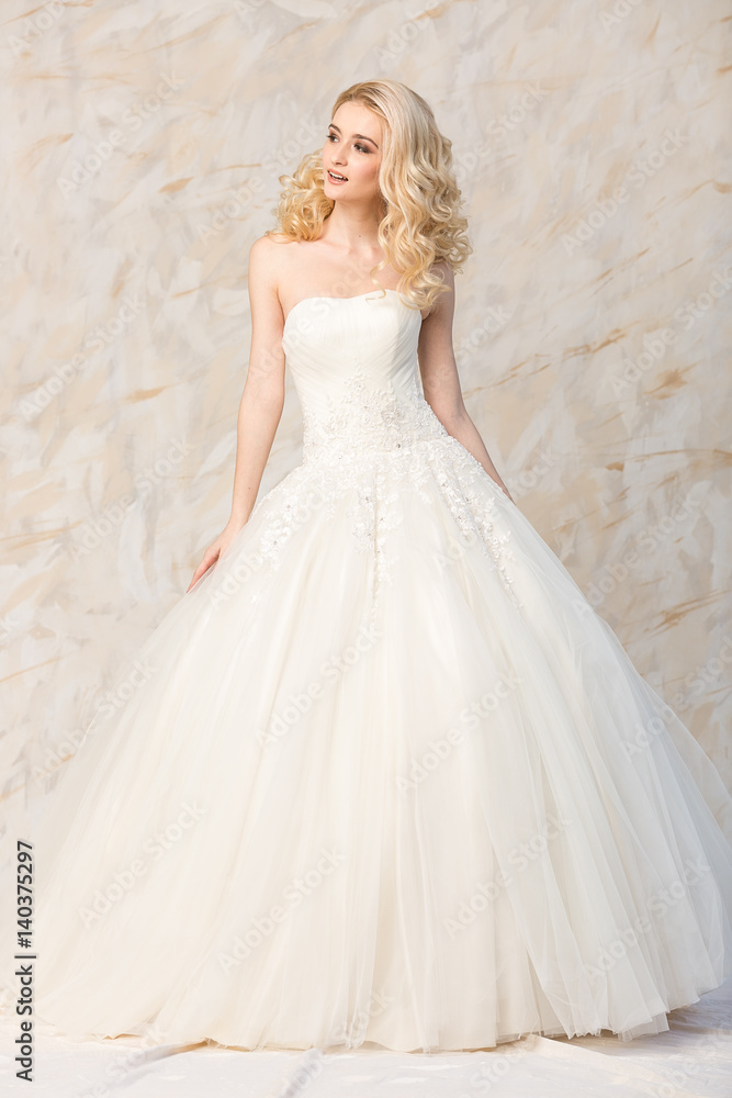 a gorgeous smiling blonde bride with curly hair in a white wedding dress to the full-length on a light background. beautiful young model with bridal hairstyle and makeup in a white magnificent dress.