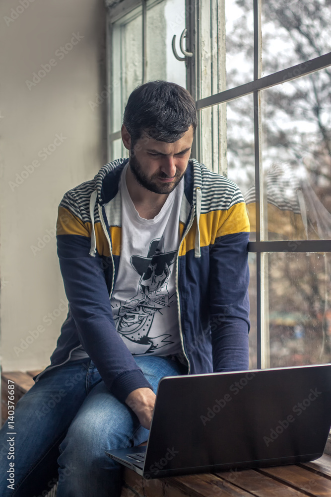 Hipster bearded man using laptop or notebook while sitting on the windowsill. Big windows on background. 