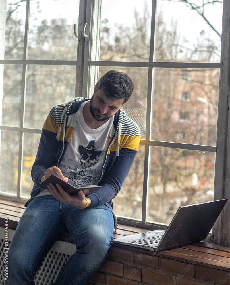 Hipster bearded man using laptop or notebook and notepad with pen while sitting on the windowsill. Big windows on background. Hipster bearded man using laptop or notebook and notepad with pen while sitting on the windowsill. Big windows on background.