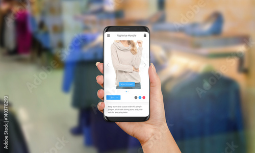 Woman buys clothes online via mobile phone. In the background is a clothing store. Modern web site or app on phone screen. Black modern phone with round edges in woman hand.