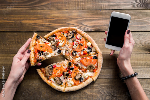 hands of a woman ordering pizza with a device over a wooden workspace table. All screen graphics are made up. photo