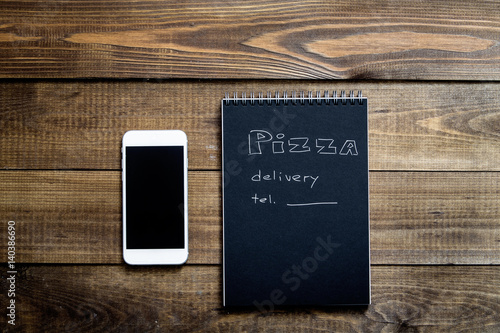 Close-up of mobile phone and notebook with the text: Pizza delivery. background wooden table. Notebook black with white text