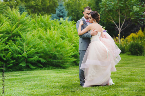Portrait of a Happy newlyweds outdoors. Back view. Bride and groom huging. Bouquet in hand. Grass, trees and bushes in the background.