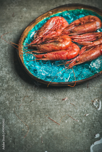 Raw uncooked red shrimps on chipped ice in turquoise blue ceramic tray over grey concrete background, copy space, vertical composition. Fresh seafood concept