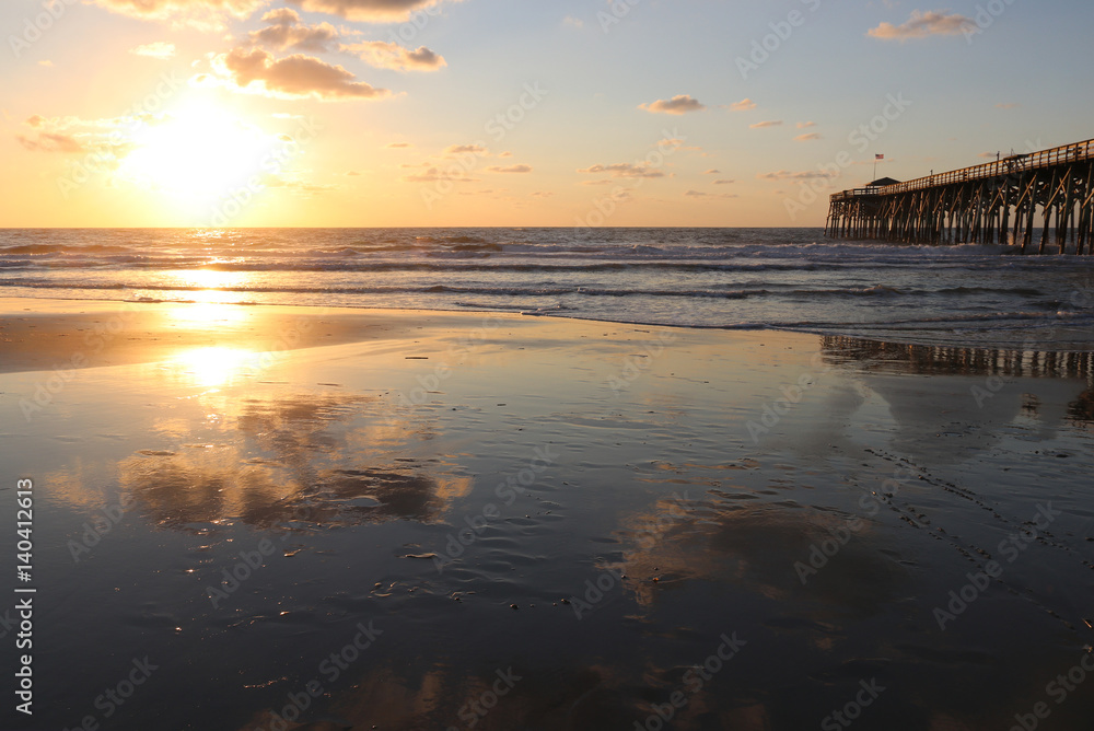 Naklejka premium Atlantic ocean sunrise background. Sunrise over the ocean. Atlantic ocean landscape with a sun and clouds reflected in shallow water and wooden pier in Myrtle Beach area, South Carolina, USA.