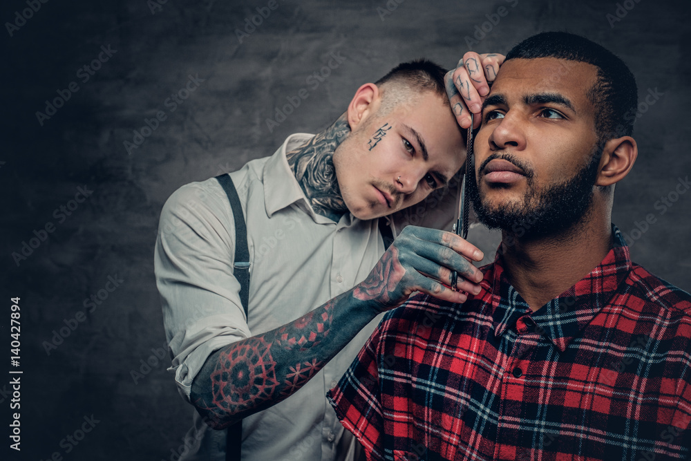 Fototapeta premium Tattooed barber cuts the beard to a Black bearded male.