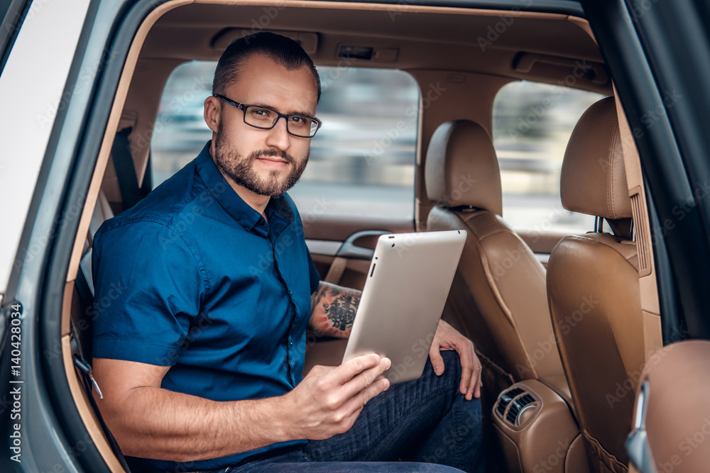 A man in eyeglasses with tattoo on his arm using portable tablet PC on a back seat of a car.