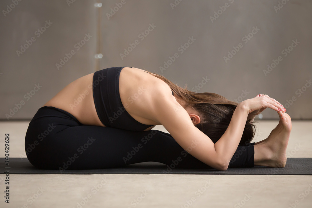 Obraz premium Young attractive sporty woman practicing yoga, stretching in paschimottanasana exercise, Seated forward bend pose, working out, wearing black sportswear, cool urban style, full length, grey studio