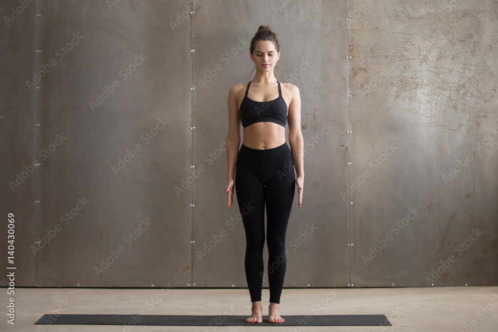 Fototapeta premium Young attractive yogi woman practicing yoga, standing in Tadasana exercise, mountain pose, working out, wearing black sportswear, cool urban style, full length, grey studio background