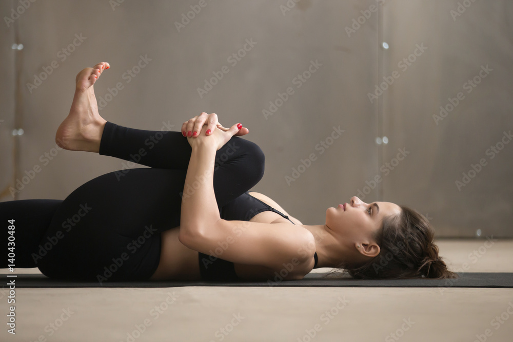 Obraz premium Young attractive sporty yogi woman practicing yoga, stretching in Knees to Chest, Apanasana exercise, working out wearing black sportswear, cool urban style, grey studio background, closeup, side view