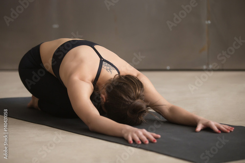 Young attractive woman practicing yoga, stretching in Child exercise, Balasana pose, working out, wearing black sportswear, cool urban style, full length, grey studio background