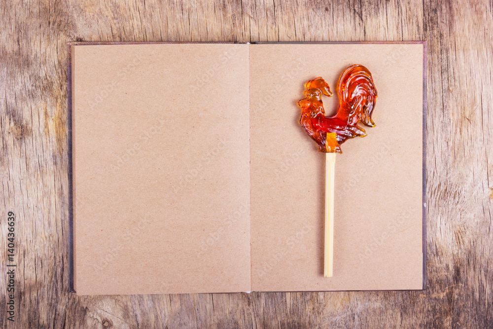 Open book with blank pages and a rooster on a stick. Lollipop on a stick in the form of a rooster on an empty sheet. Copy space. Open book with blank pages and a rooster on a stick. Lollipop on a stick in the form of a rooster on an empty sheet. Copy space.