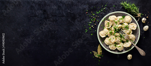 Traditional russian pelmeni, ravioli, dumplings with meat on black concrete background. Parsley, quail eggs, pepper, rosemary, bay leaf and spices. Top view. Copyspace. Banner.