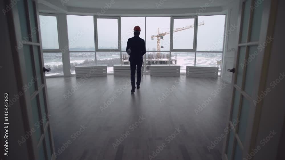 Man wearing in suit and helmet holding documents with architectural plan. rear back view businessman looking through the windows on the construction site with high-rise Man wearing in suit and helmet holding documents with architectural plan. rear back view businessman looking through the windows on the construction site with high-rise