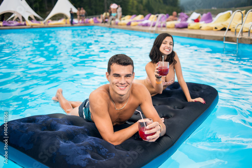 Handsome man with cocktail lying on an inflatable mattress in pool with happy female out of focus next to him