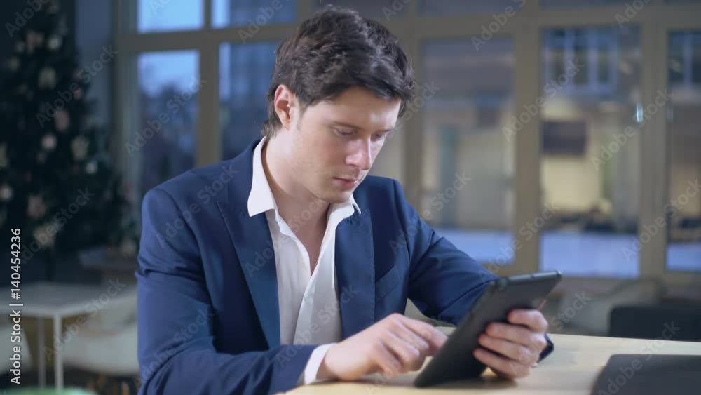 Portrait handsome adult man using touch screen tablet. Caucasian young male surfing internet or chatting in application. Businessman wearing in casual elegant jacket sitting in room with big windows Portrait handsome adult man using touch screen tablet. Caucasian young male surfing internet or chatting in application. Businessman wearing in casual elegant jacket sitting in room with big windows