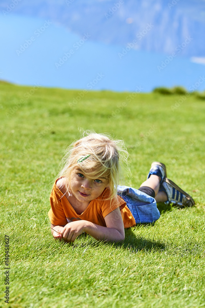 Fototapeta premium Child blond girl lying on grass over the lake Lago di Garda on the top of hill Monte Baldo, Italy