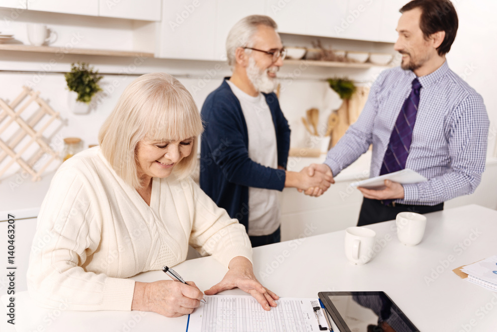 Retired woman signing contract while her husband greeting the notary Retired woman signing contract while her husband greeting the notary