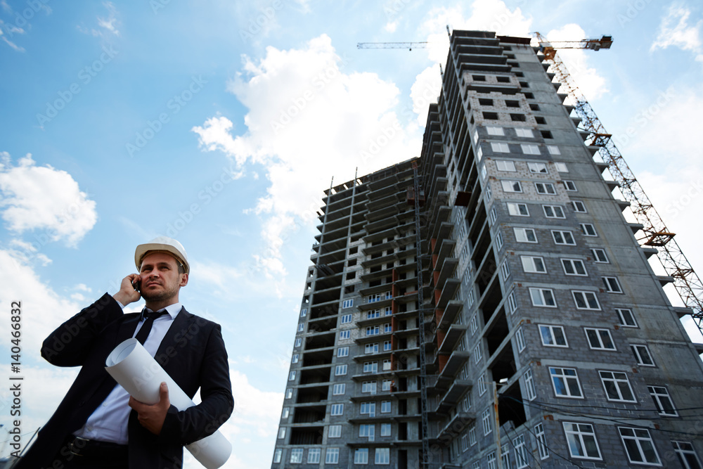 Elegant investor in helmet talking on mobile phone by unfinished building Elegant investor in helmet talking on mobile phone by unfinished building