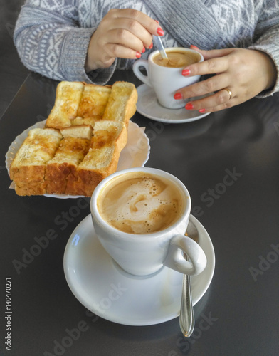 Hands of woman holding cup of coffee with milk and toast with butter