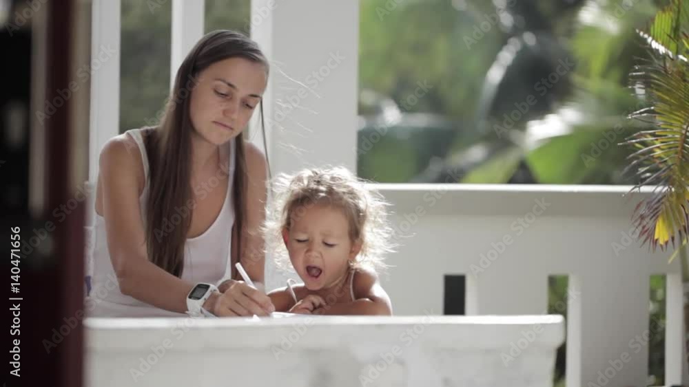 Young mother with daughter drawing at balcony with palm trees on the background