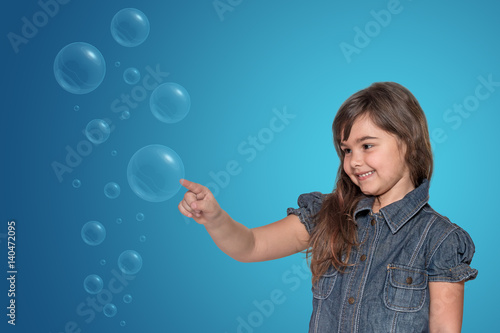 Smiling little girl is touching the transparent bubbles. All is on on the blue gradient background with light rays. 