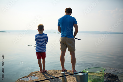 Back view portrait of muscular well-built man adjusting fishing rod while standing on lake with his little son, sharing hobby on sunny summer day