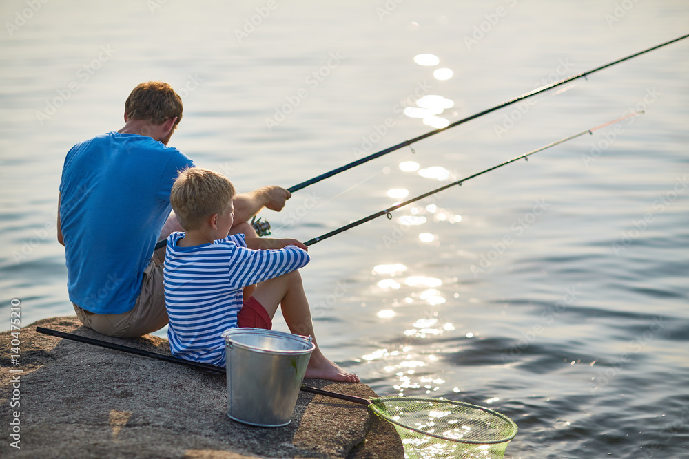 Back view portrait of adult man and teenage boy sitting together fishing with rods in calm glittering waters of blue lake on sunny summer day Back view portrait of adult man and teenage boy sitting together fishing with rods in calm glittering waters of blue lake on sunny summer day