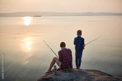 Back view portrait of adult man and teenage boy sitting together on rocks fishing with rods in calm waters with landscape of setting sun, both wearing checkered shirts