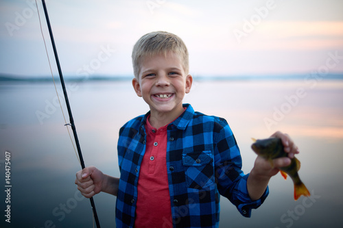Portrait of blond happy boy smiling looking at camera holding fishing rod and single perch against calm blue lake background at dusk
