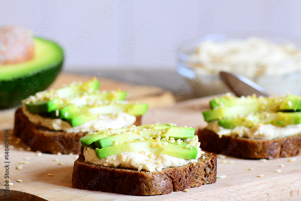 Hummus avocado sandwiches on a wooden board. Sandwiches made with rye bread, fresh avocado slices, hummus and roasted sesame seeds. Healthy eating for the whole family