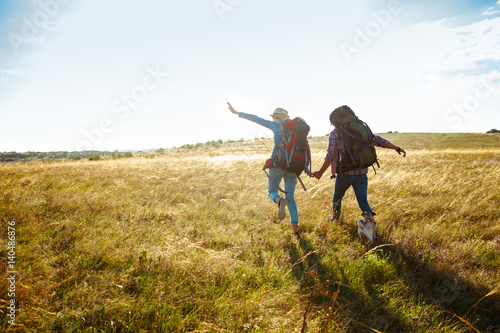 Young couple of travelers walking in field with pug dog.