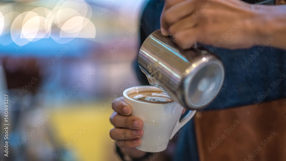 Barista is pouring milk froth in order to make latte art on a hot coffee cup.