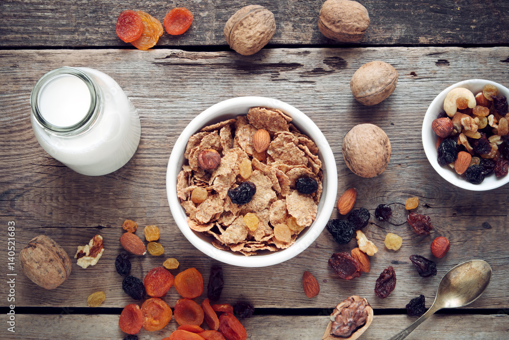 Ingredients for healthy breakfast: cereal wheat flakes in bowl, dried fruits, muesli and milk bottle on wooden table. Top view, flat lay. Ingredients for healthy breakfast: cereal wheat flakes in bowl, dried fruits, muesli and milk bottle on wooden table. Top view, flat lay.