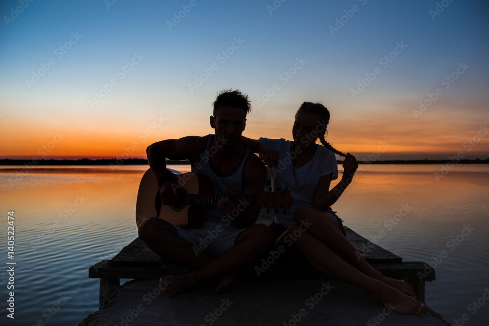 Silhouettes of young beautiful couple resting rejoicing at sunrise near lake.