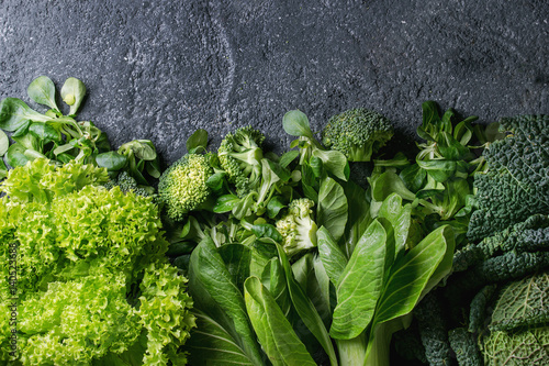 Variety of raw green vegetables salads, lettuce, bok choy, corn, broccoli, savoy cabbage as frame over black stone texture background. Top view, space for text