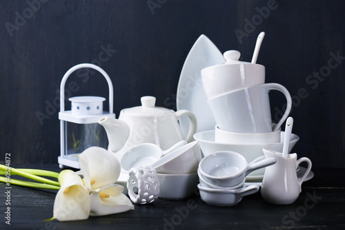Set of new white dishes with teapot, tea cups, and plates on a black wooden table with fresh callas