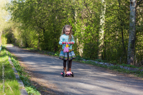 Little child learning to ride a scooter in a city park on sunny summer day. Kids play outdoors.